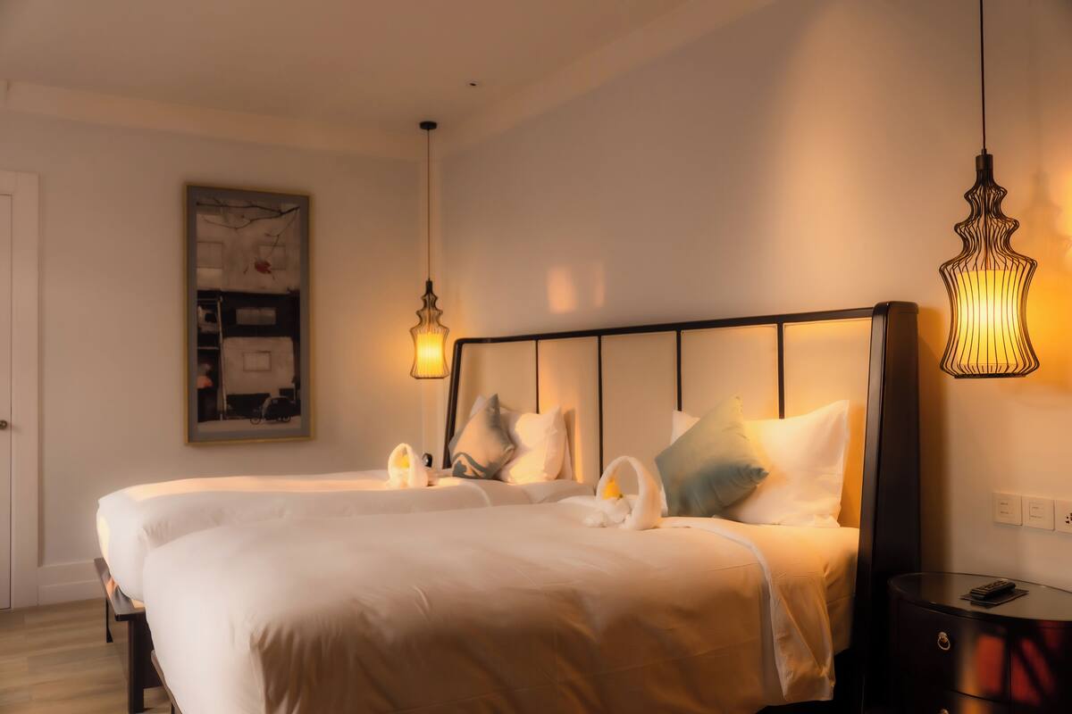 Hotellihuone, jossa on kaksi sänkyä, suuri taulu seinällä ja moderni riippuvalaisin.