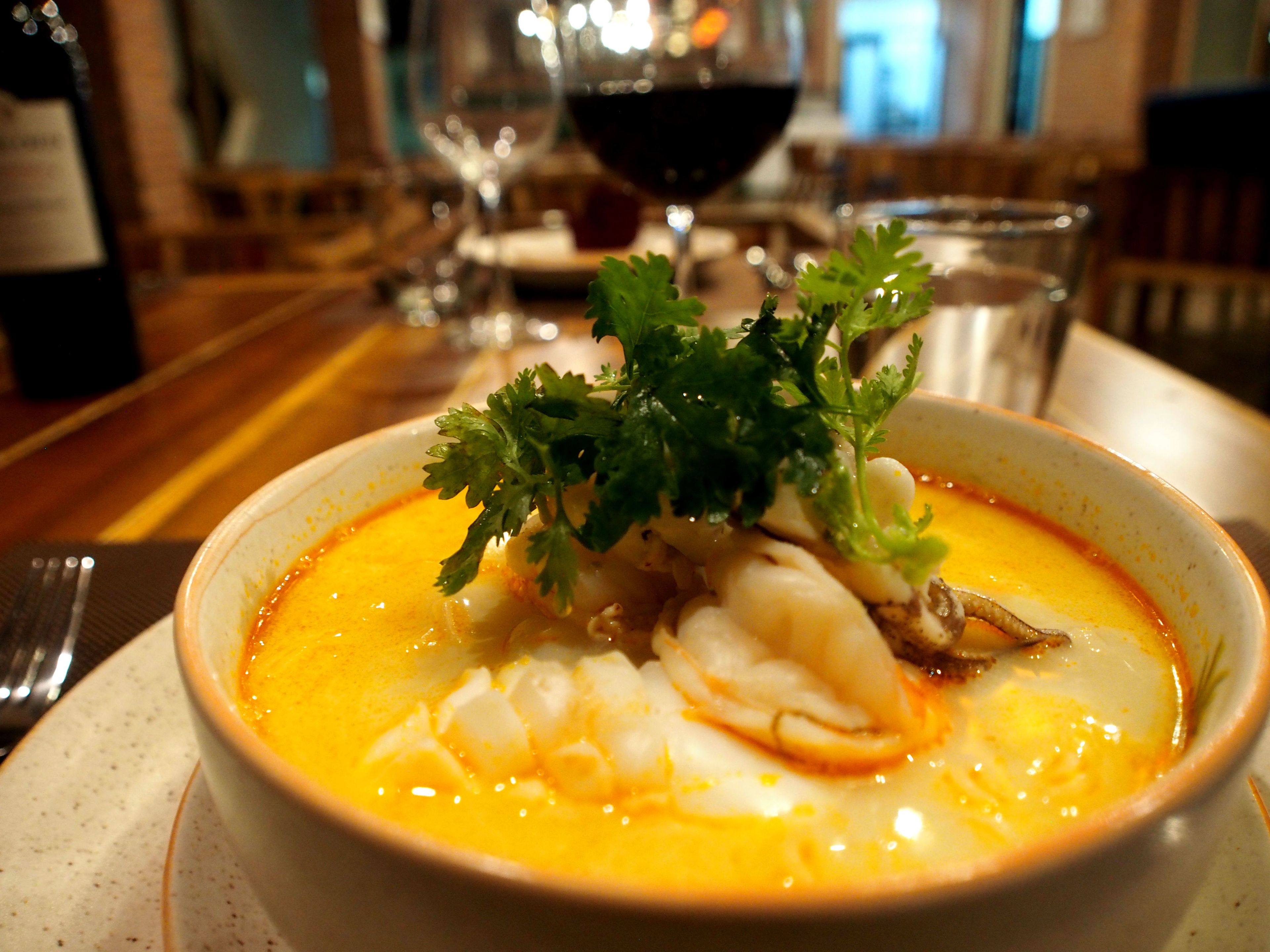 A bowl of seafood soup garnished with parsley, served on a plate with a fork and knife beside it.