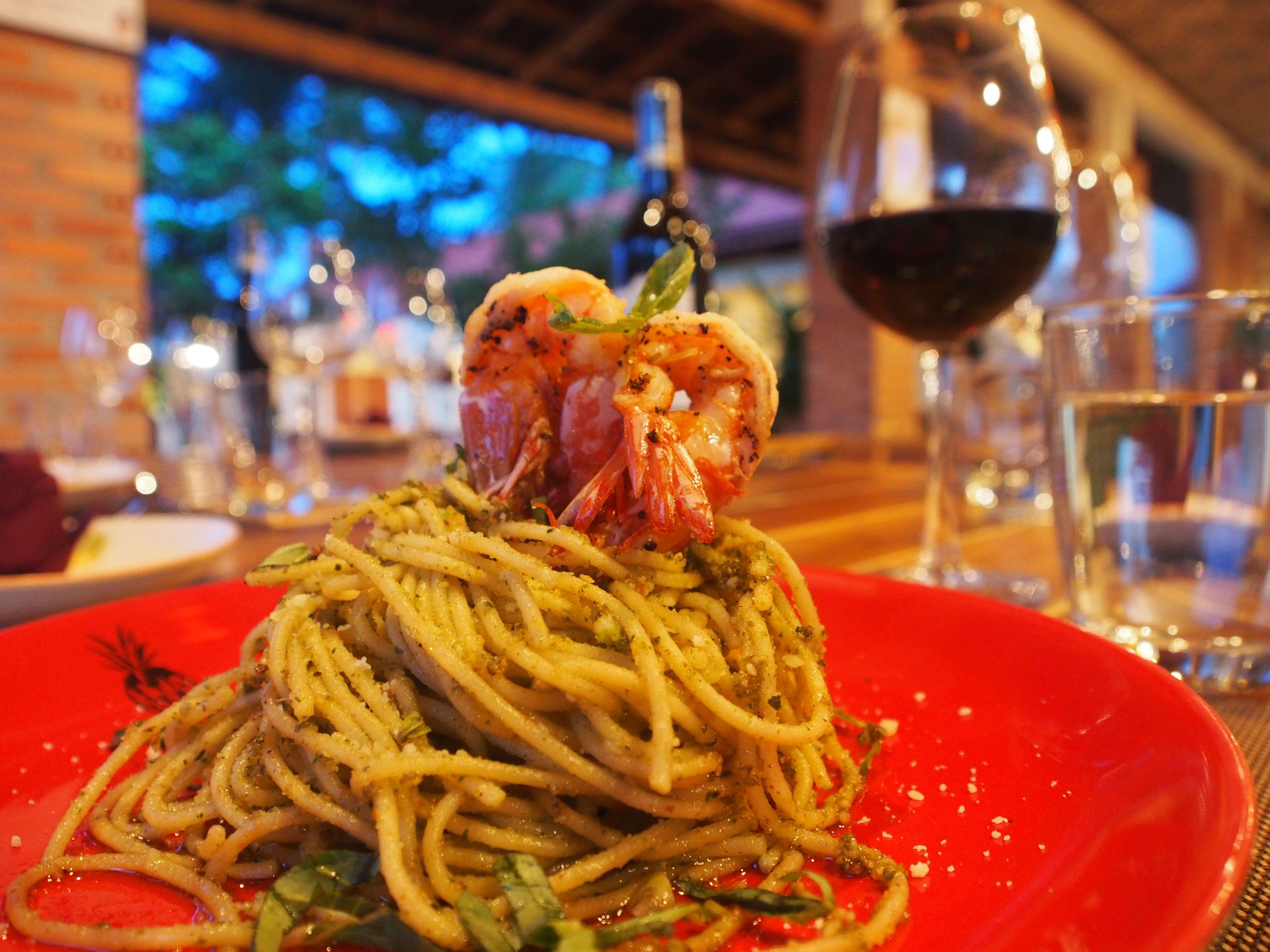 Eine Teller Spaghetti mit einem Garnelen-Spieß darauf, ein Glas Rotwein und ein Glas Wasser auf einem Tisch.
