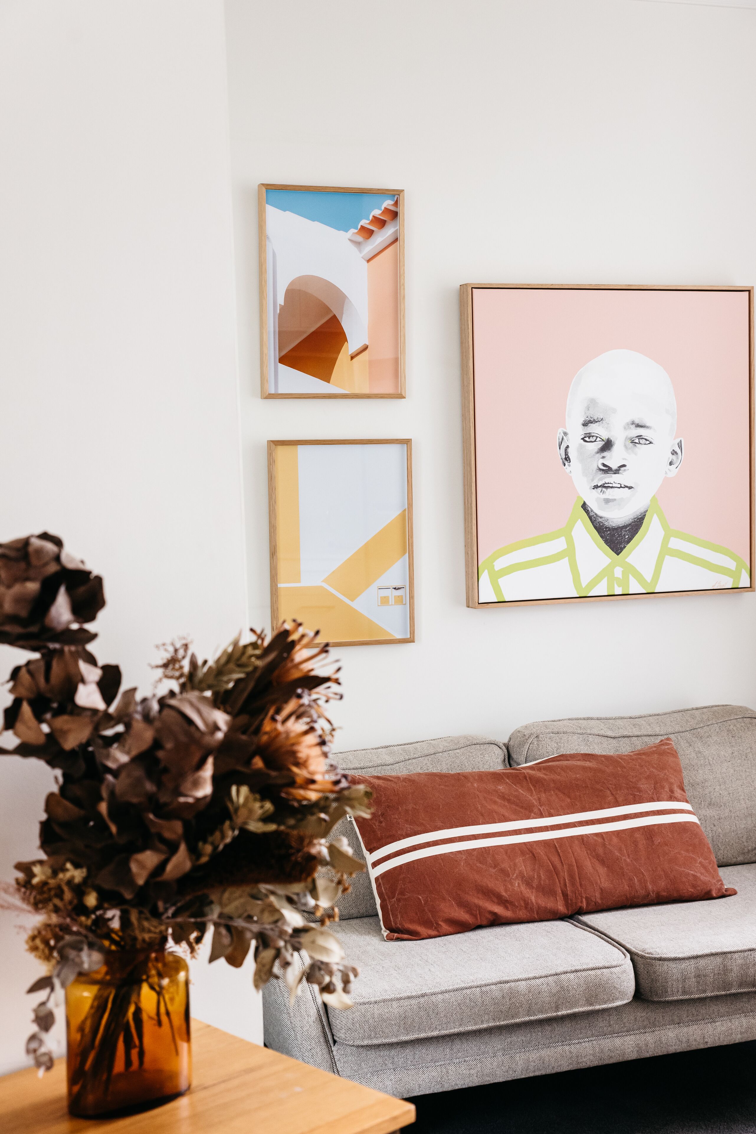 A living room with a grey sofa, a brown cushion, and a vase with dried flowers. There are three framed artworks on the wall.