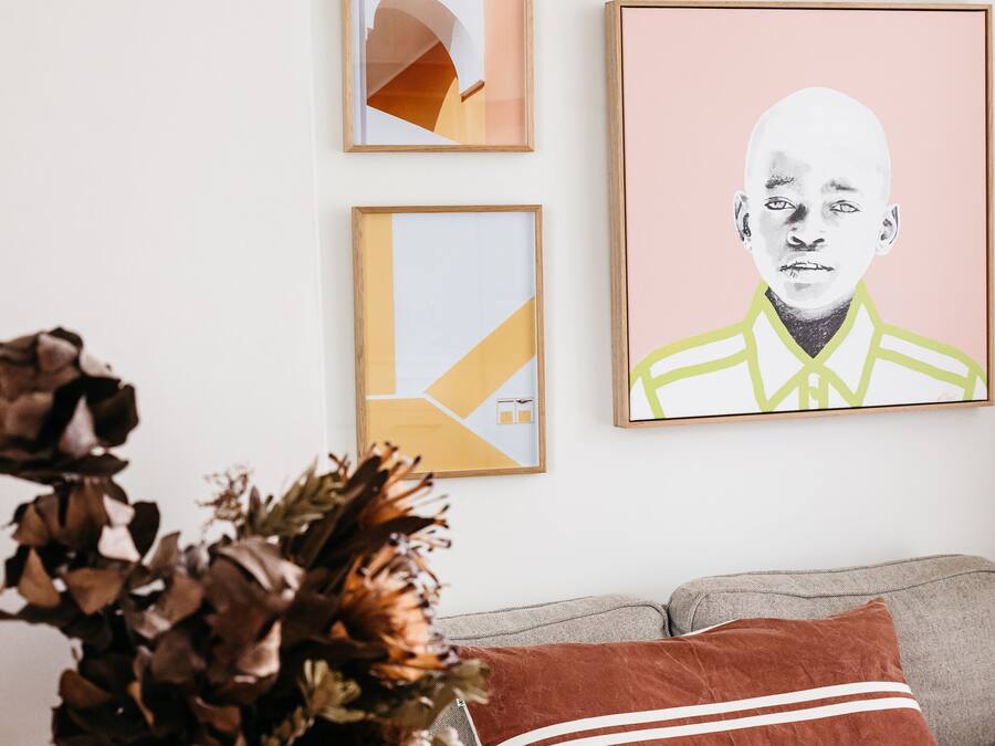 A living room with a grey sofa, a brown cushion, and a vase with dried flowers. There are three framed artworks on the wall.