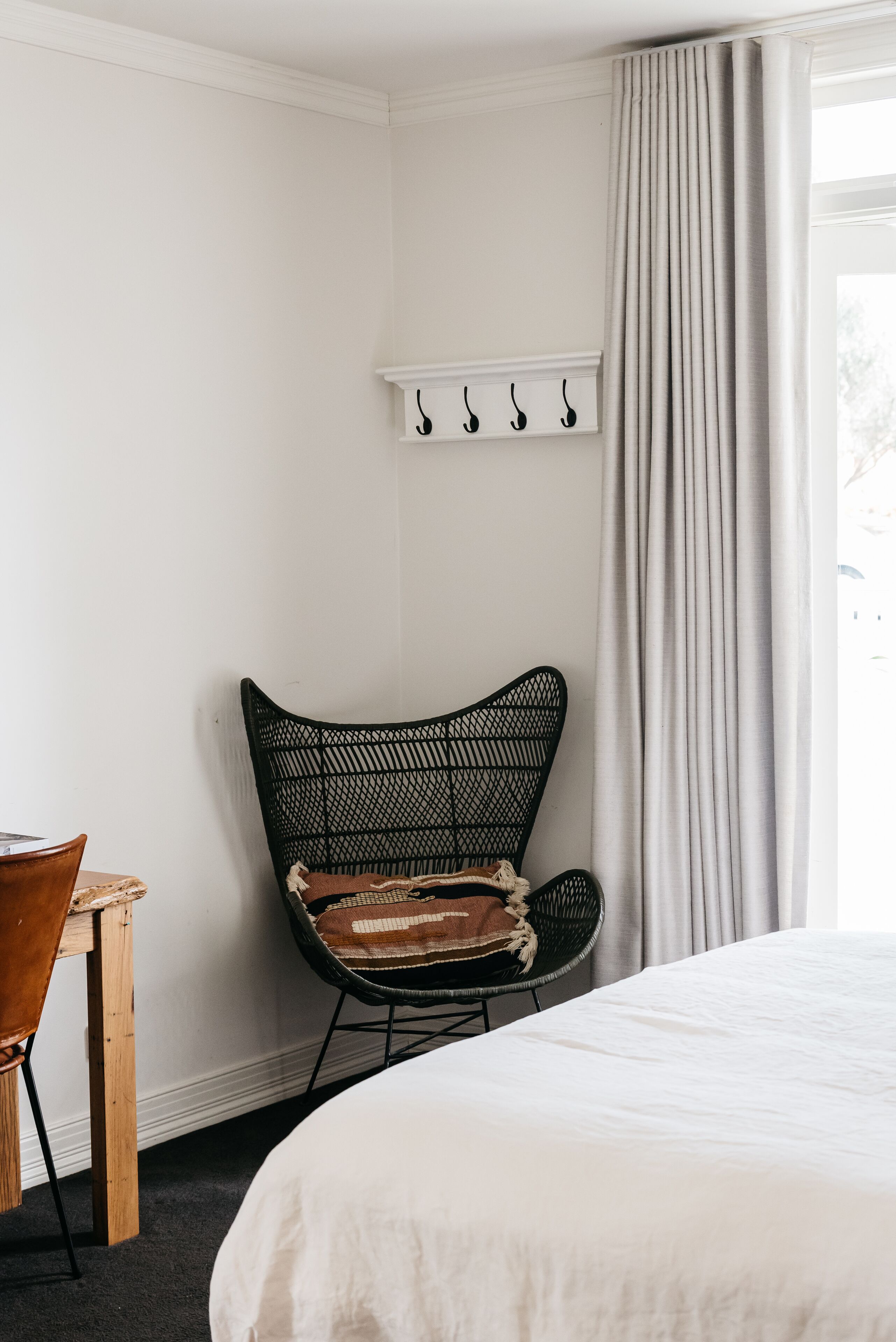 Una habitación con cama, una silla, una mesita, un estante con ganchos y una ventana con cortinas.