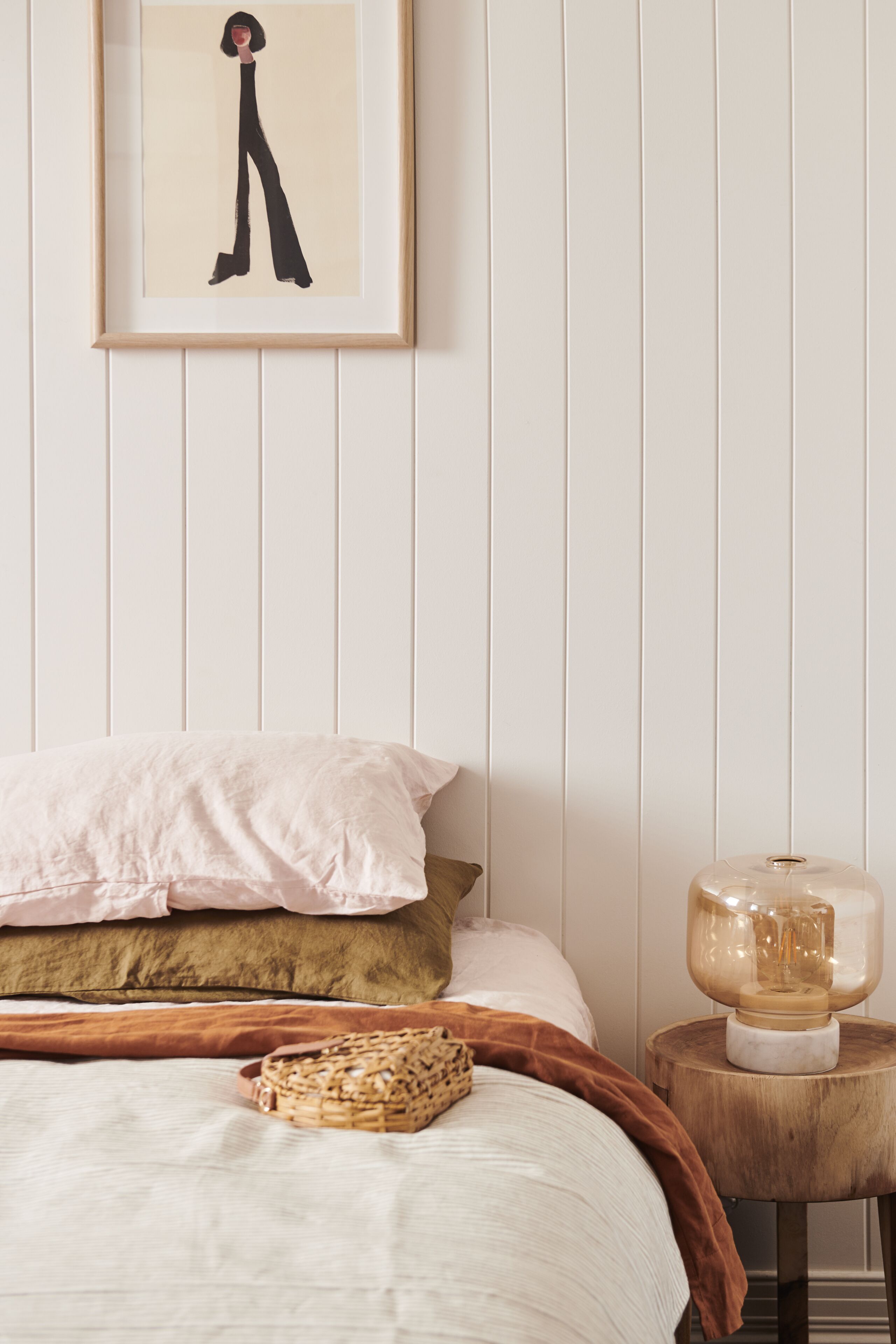 A neatly arranged bedroom with a bed, pillows, a bedside table with a lamp, and a framed artwork on the wall.
