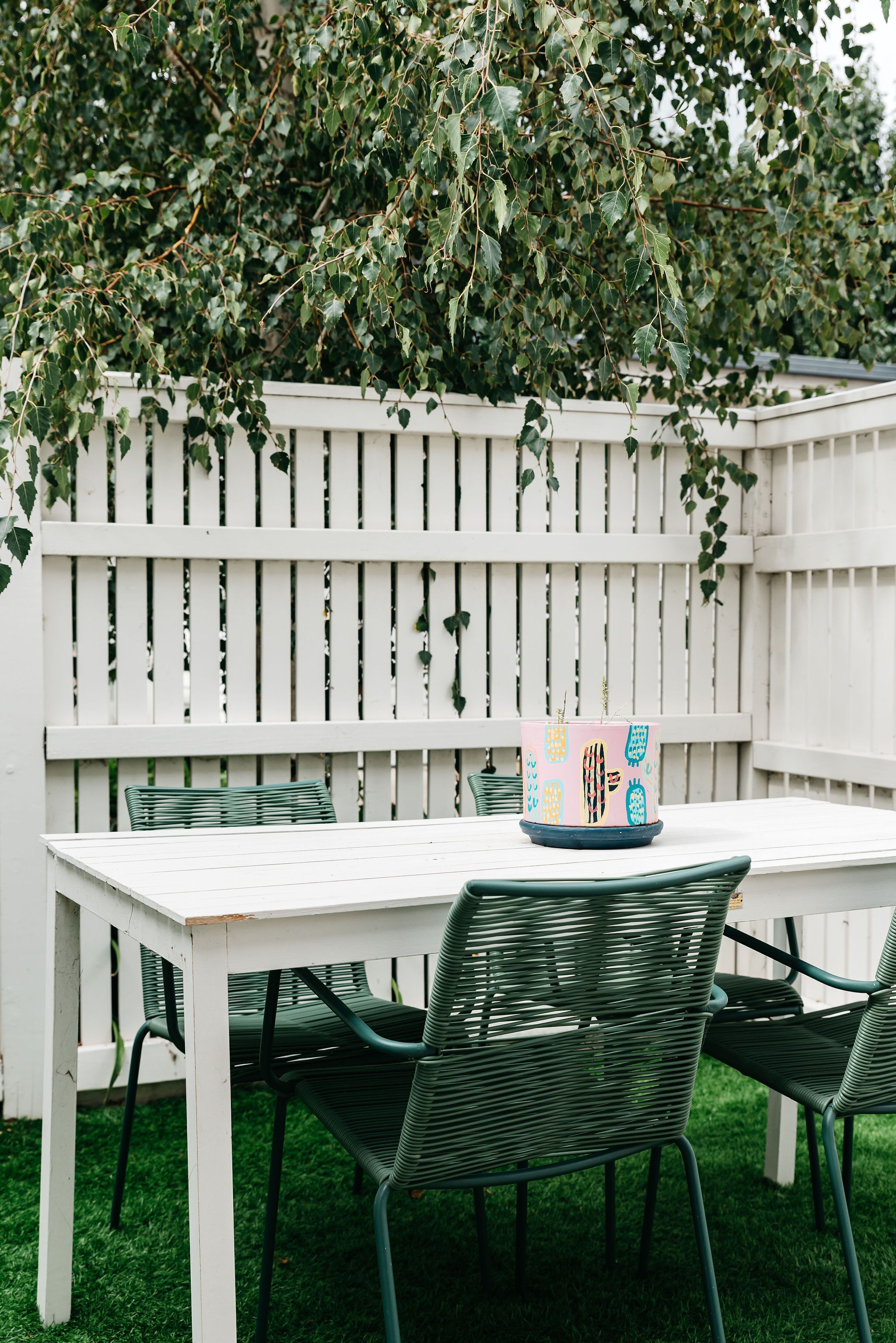 A white wooden table with four green chairs, set against a white fence with climbing ivy.