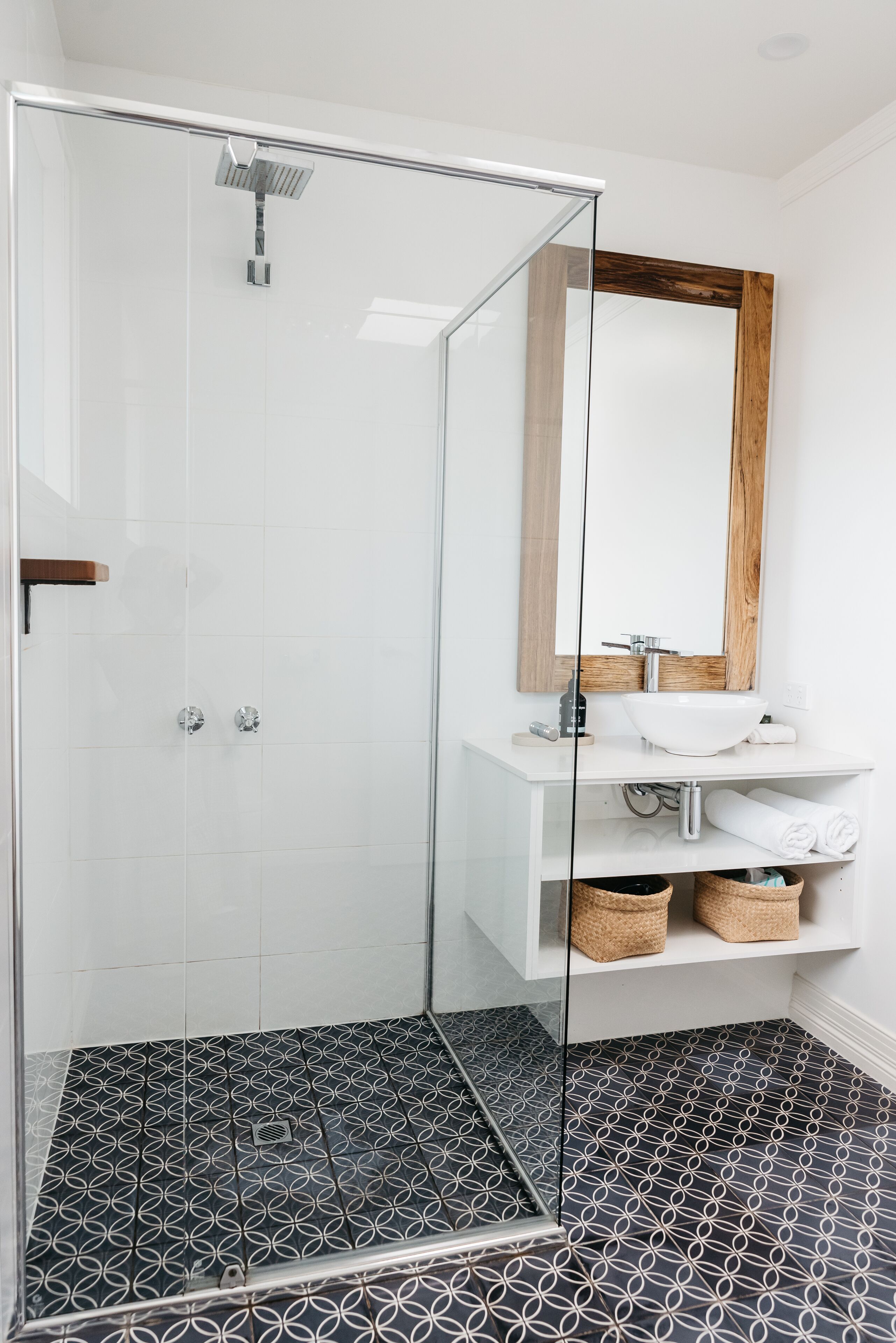 A modern bathroom with a glass-enclosed shower, white tiles, a wooden-framed mirror, and a white sink with a towel rack.