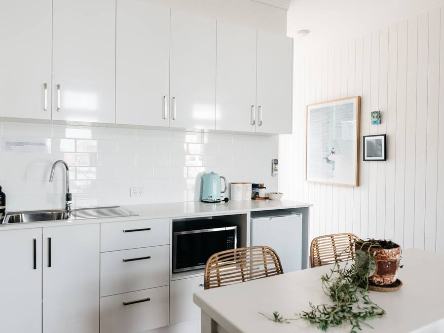 Una cucina moderna con mobili bianchi, un lavello in acciaio inossidabile, un forno a microonde e una macchina da caffè. Ci sono due sedie in vimini e un tavolo con una pianta in vaso.