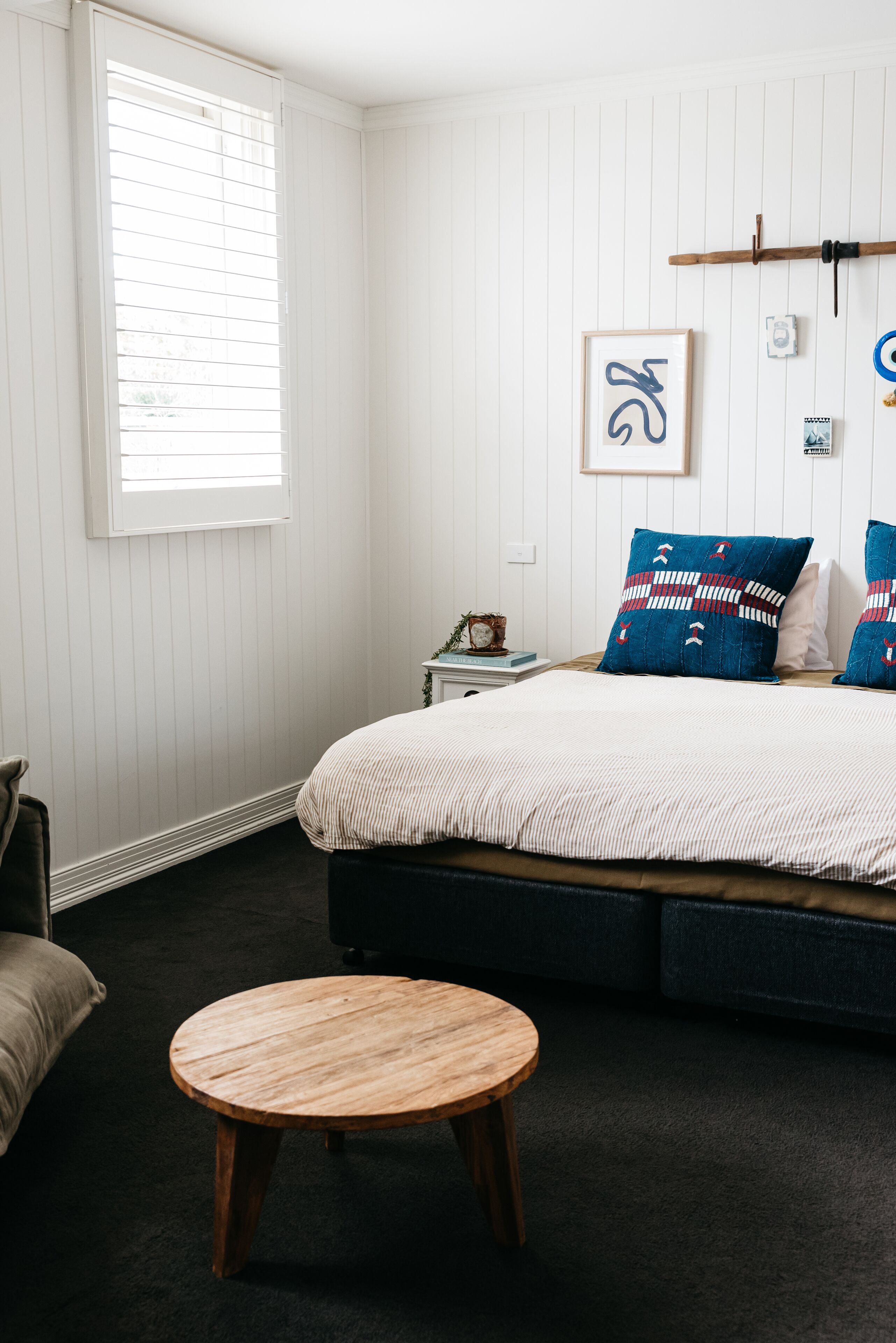 Une chambre à coucher avec un lit, une table ronde en bois, une fenêtre avec des stores et un tableau accroché au mur.