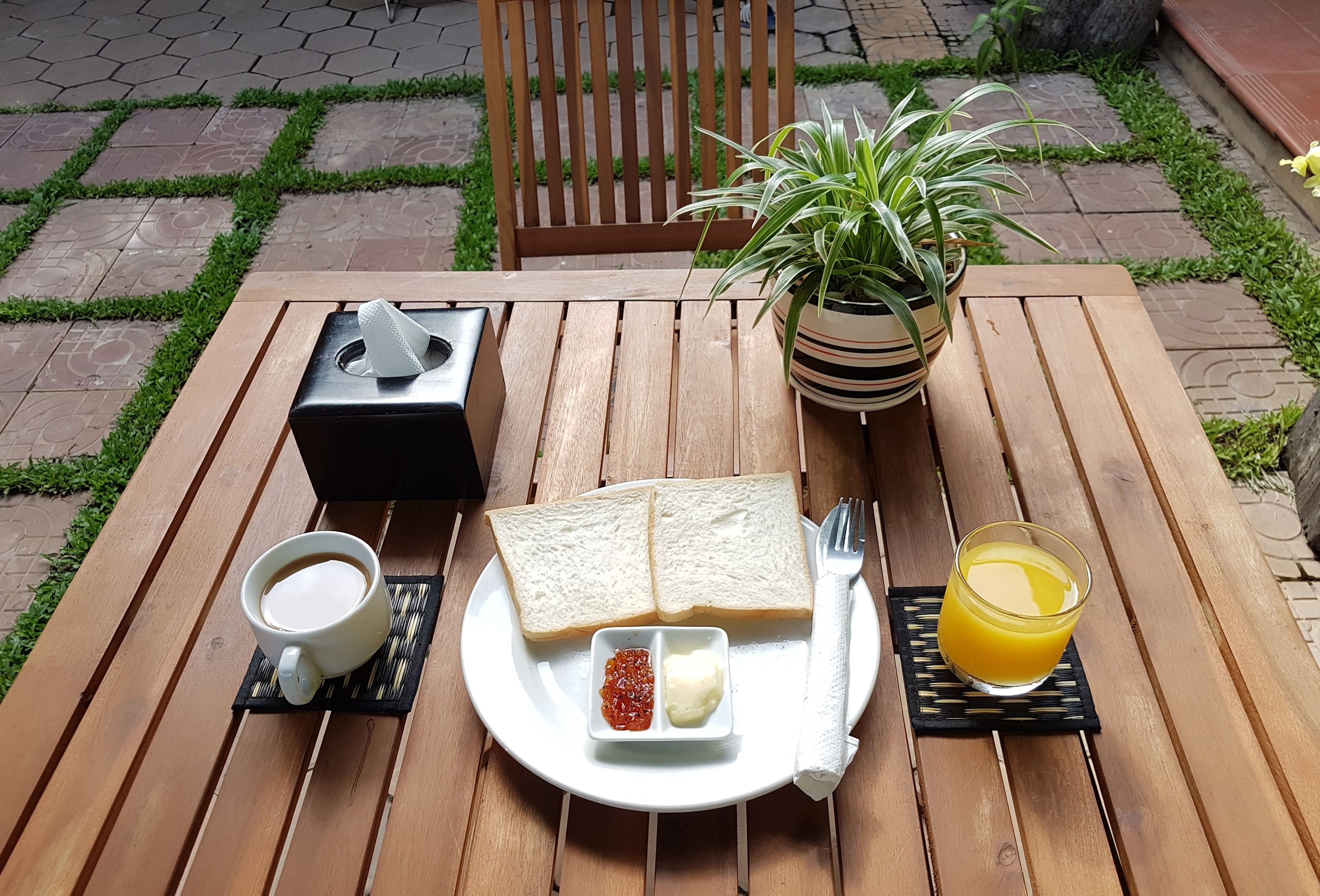 Une table en bois sur laquelle se trouvent une assiette de toasts, du beurre, de la confiture, une tasse de café et un verre de jus d’orange.