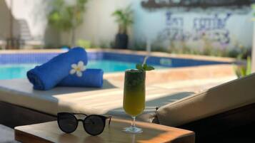 A poolside setting with a lounge chair, a rolled-up towel, a glass of a yellow beverage, and a pair of sunglasses on a small table.