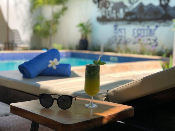 Une scène au bord de la piscine, avec un fauteuil de relaxation, une serviette enroulée, un verre d’une boisson jaune et une paire de lunettes de soleil posées sur une petite table.