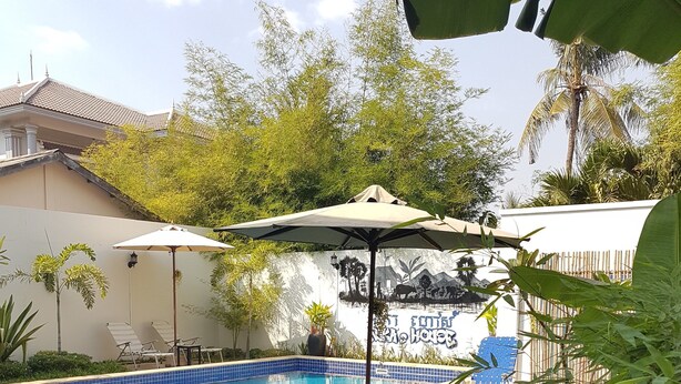 A swimming pool with lounge chairs and umbrellas, surrounded by greenery and a mural. - Nika's House (Siem Reap)