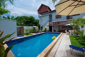 A swimming pool with a blue tiled edge, surrounded by a garden with various plants and a white fence. - Nika's House (Siem Reap)