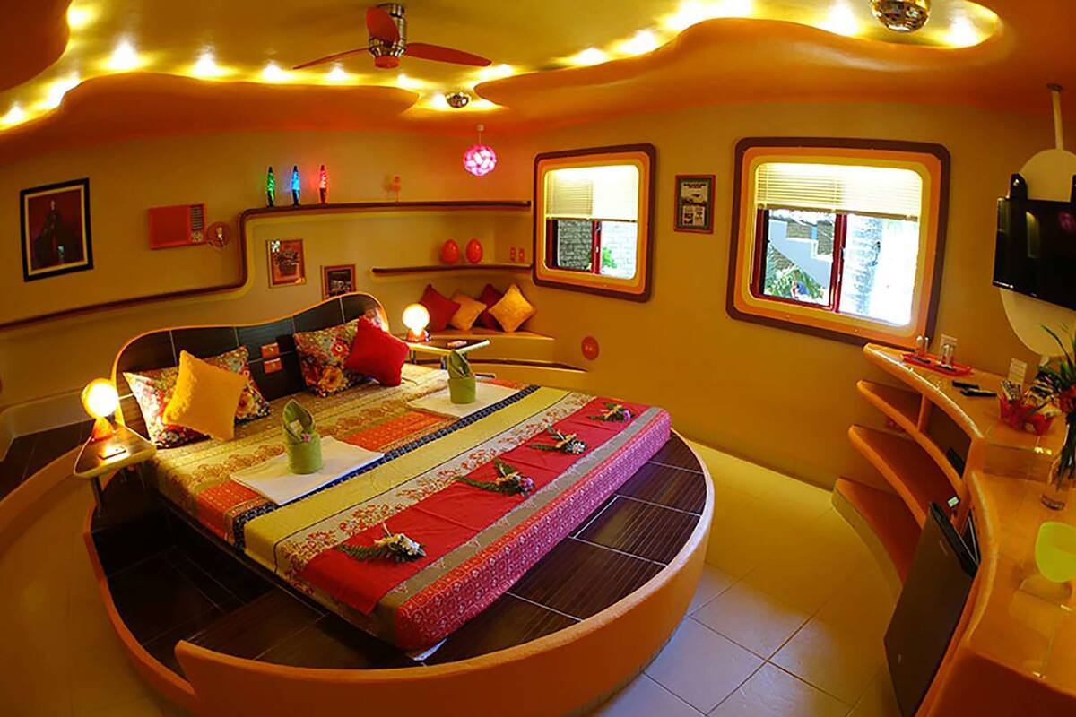 A circular bed with a red and yellow bedspread, a ceiling fan, and two windows with blinds.