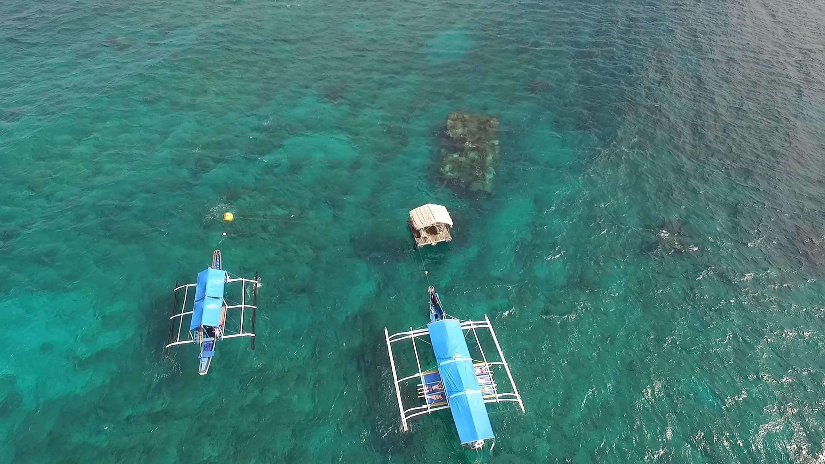 An aerial view of a clear blue sea with two floating structures, one with a blue canopy and the other with a thatched roof, connected by a rope.