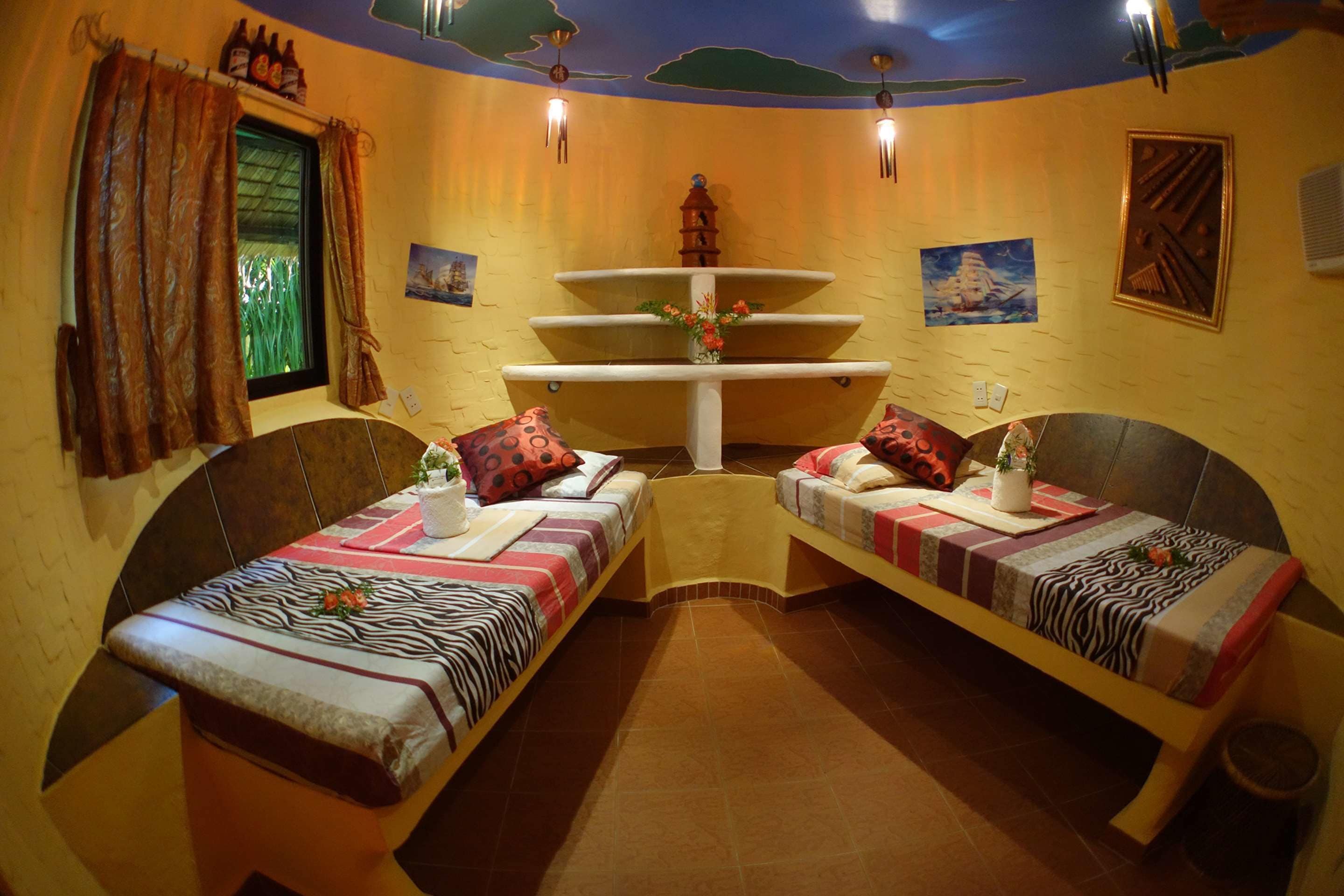 A room with two single beds, a small table, a window with curtains, a decorative wall piece, and a ceiling fan.