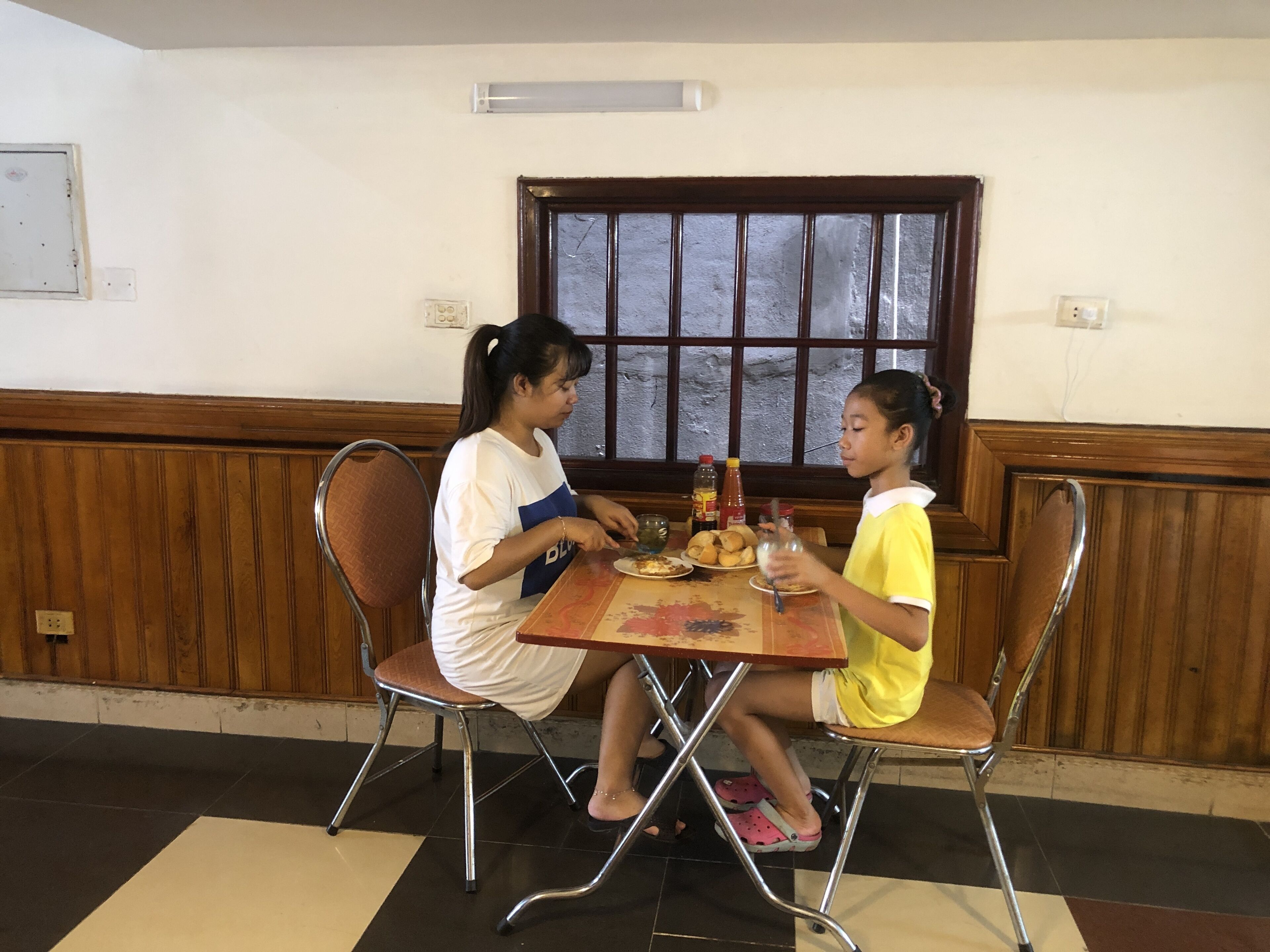 Two people seated at a table with food and drinks, in a room with wooden paneling and a window.