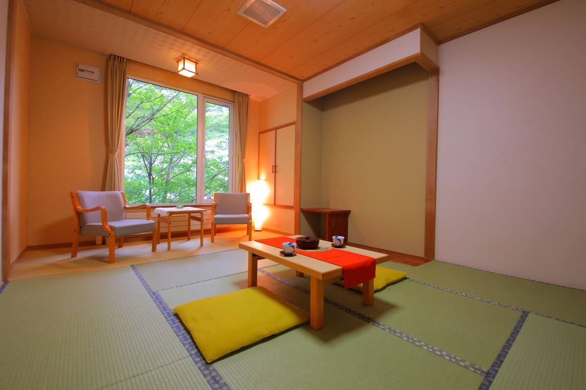 A traditional Japanese room with tatami flooring, a low wooden table, and a view of trees through the window.