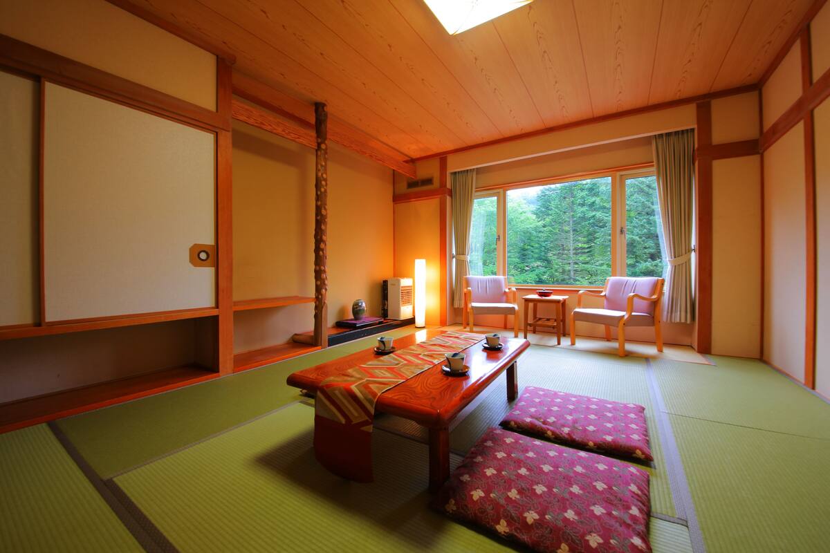 A traditional Japanese room with tatami flooring, a low wooden table with cushions, a sliding door, and a view of a forest through a large window.