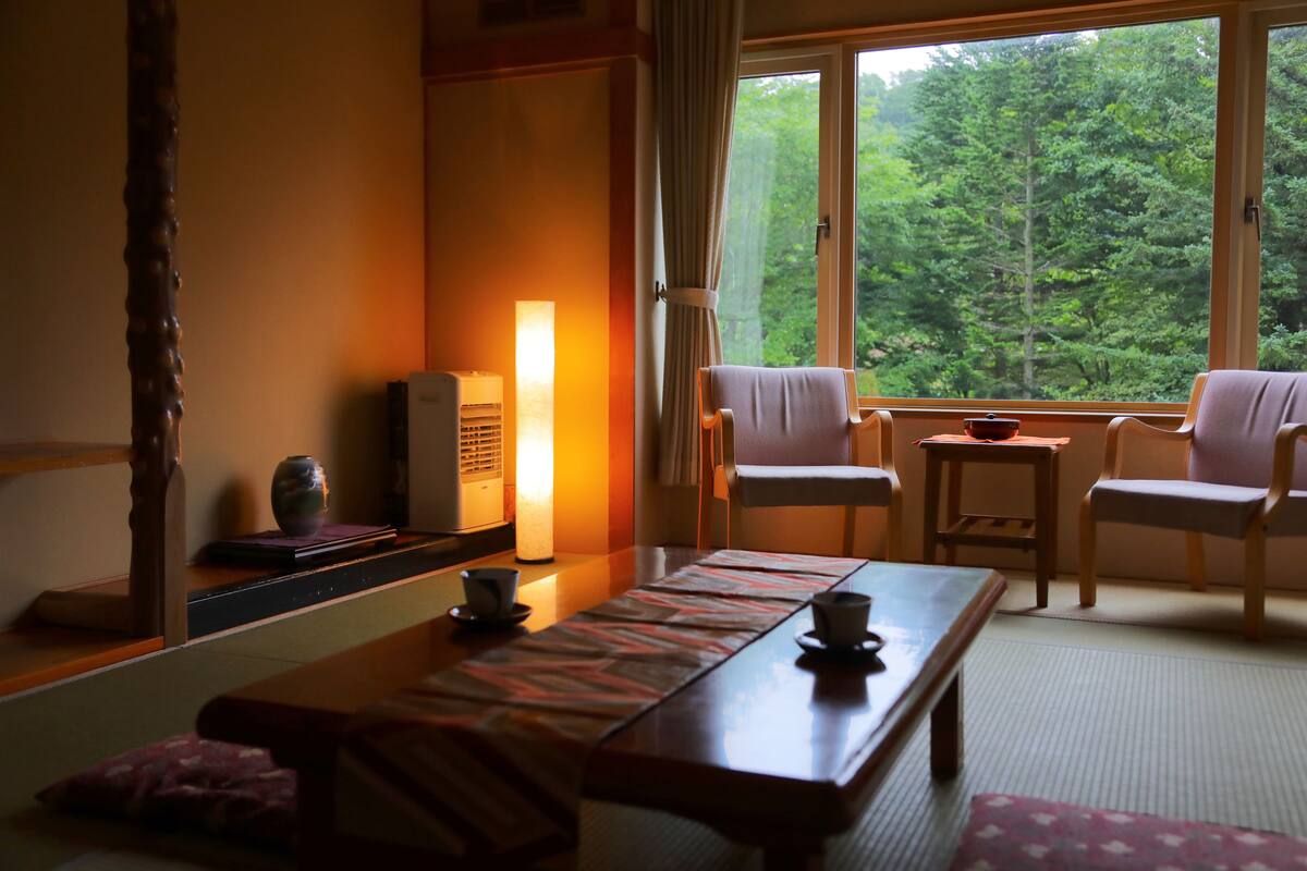 A traditional Japanese-style room with tatami flooring, a low table with a tea set, and a view of a forest through large windows.