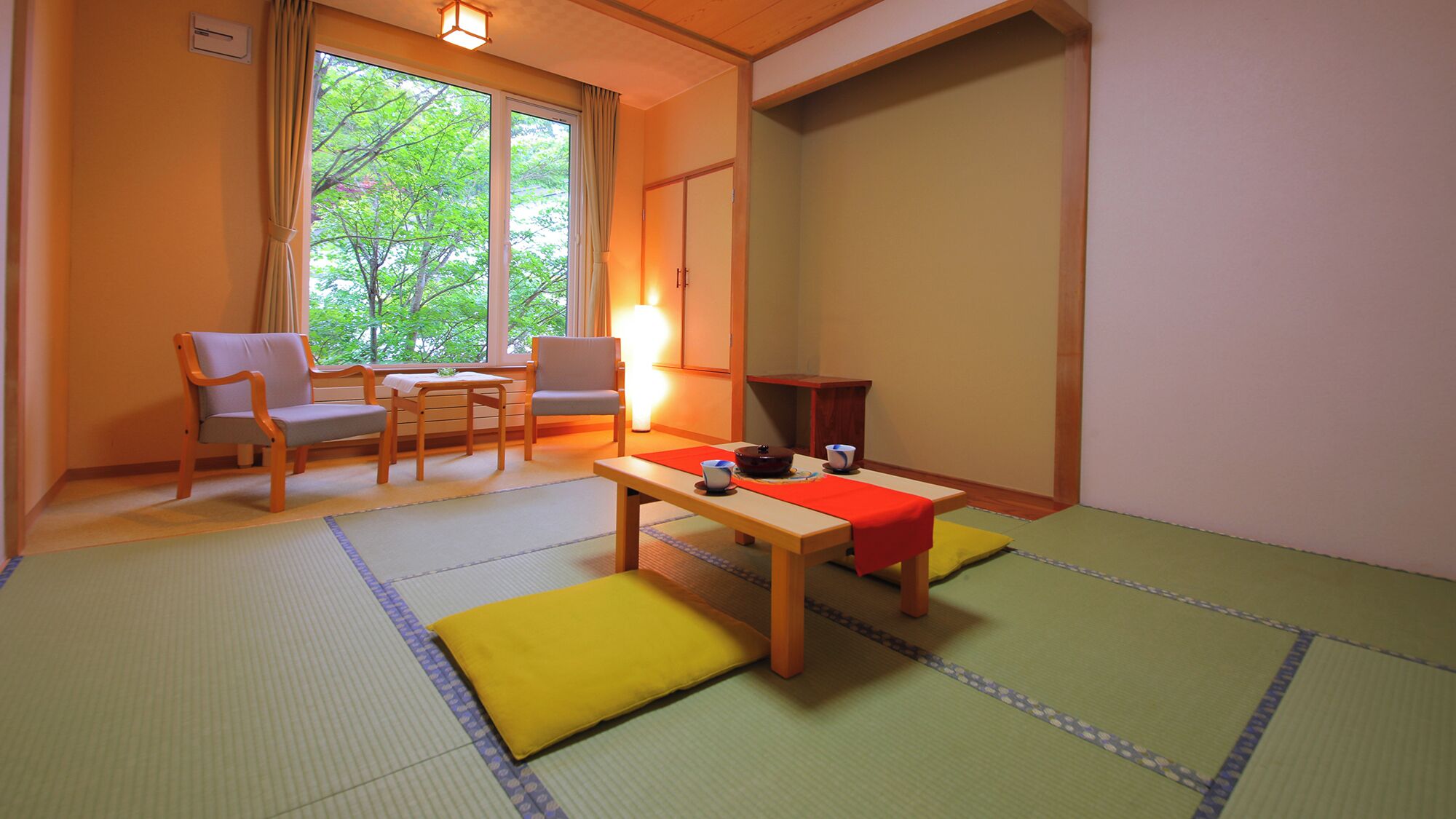 Ein traditioneller japanischer Raum mit Tatami-Bodenbelag, einem niedrigen Tisch mit roher Matte, zwei Stühlen, einem Fenster mit Vorhängen und einer Schiebetür.
