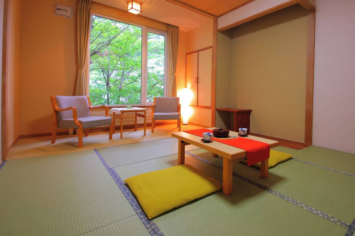 A traditional Japanese room with tatami flooring, a low table with a red mat, two chairs, a window with curtains, and a sliding door.