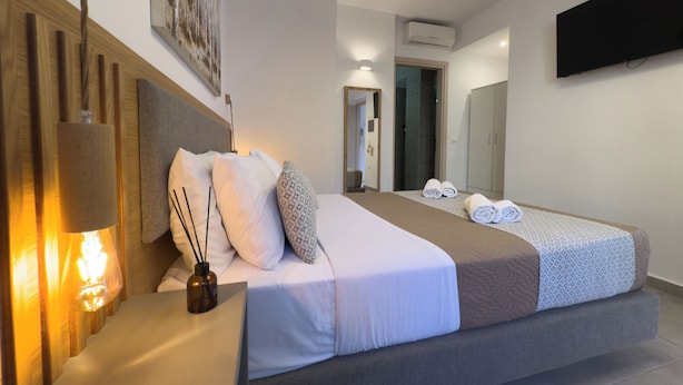 Habitación de hotel moderna con una cama grande, una mesita de noche con lámpara, un televisor montado en la pared y una zona de ducha visible a través de un vidrio.
