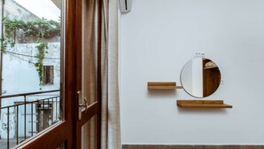 Una habitación con puerta de madera, ventana con vista a un balcón, espejo redondo en la pared y una pequeña estantería.