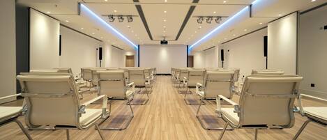 Un salón de conferencias con filas de sillas, suelo de madera y techo blanco con iluminación empotrada.