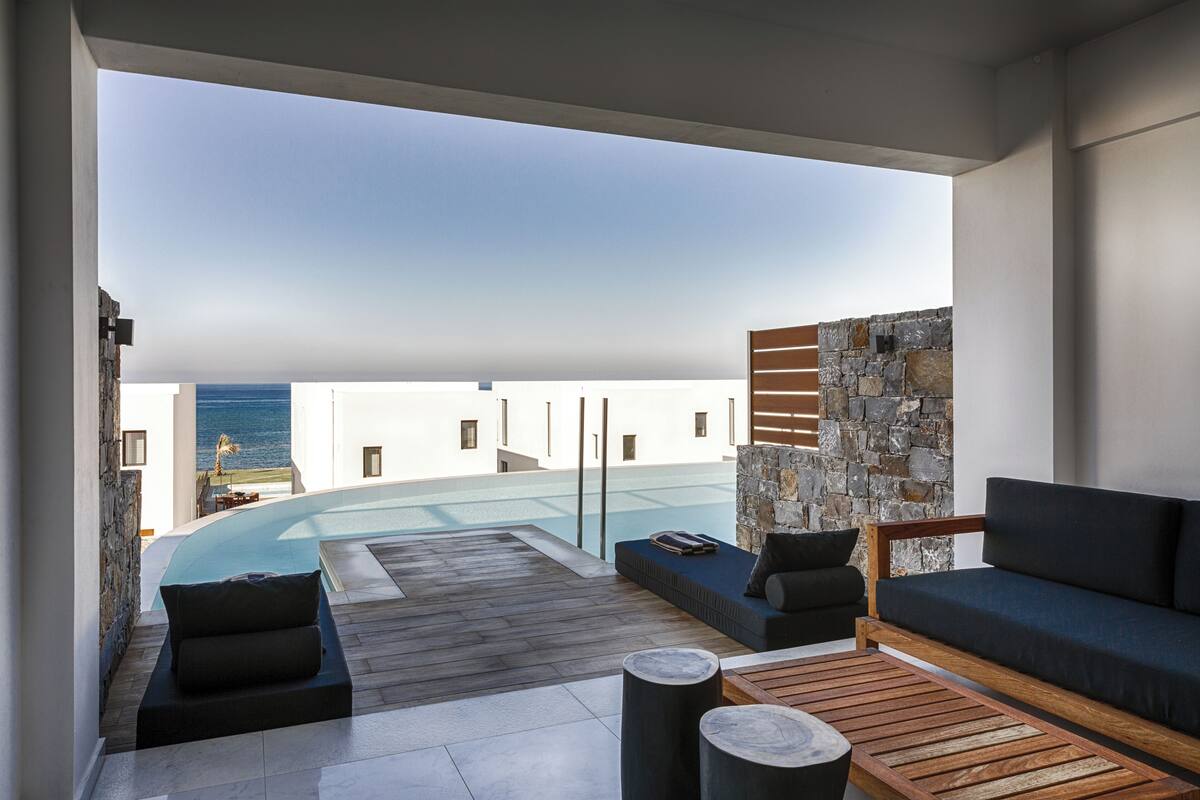 A modern outdoor lounge area with a pool, stone walls, and a view of the sea.