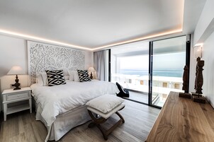 A modern bedroom with a large bed, a nightstand, a wooden bench, and a view of the ocean through floor-to-ceiling windows.