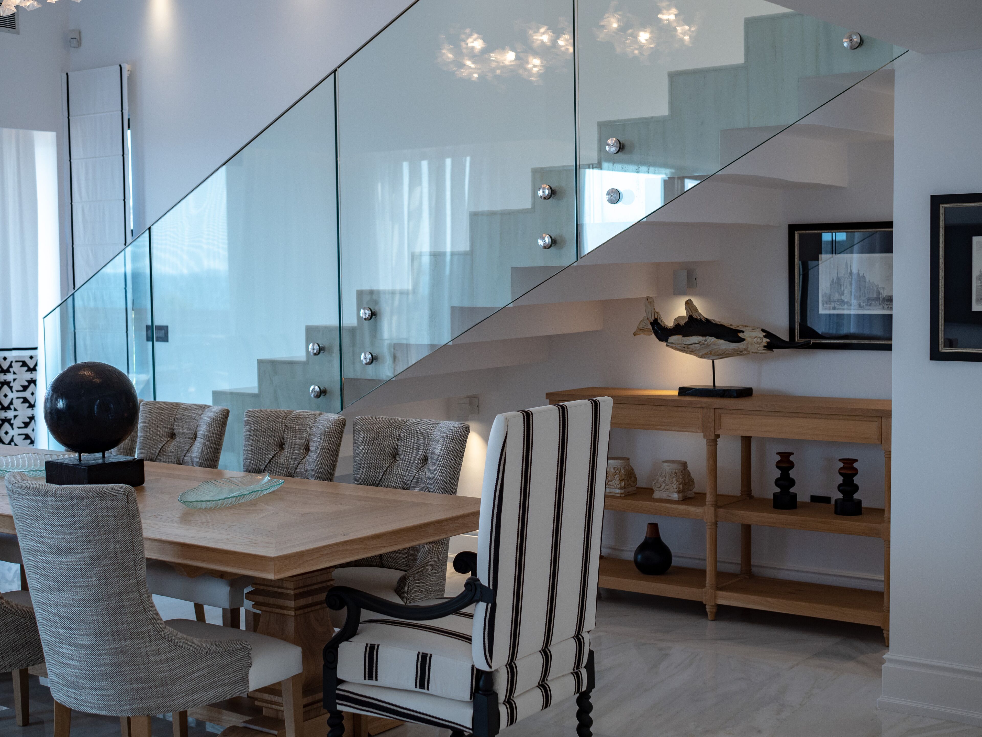 A modern interior with a dining area featuring a wooden table and upholstered chairs, a staircase with glass railings, and a wall-mounted sculpture.