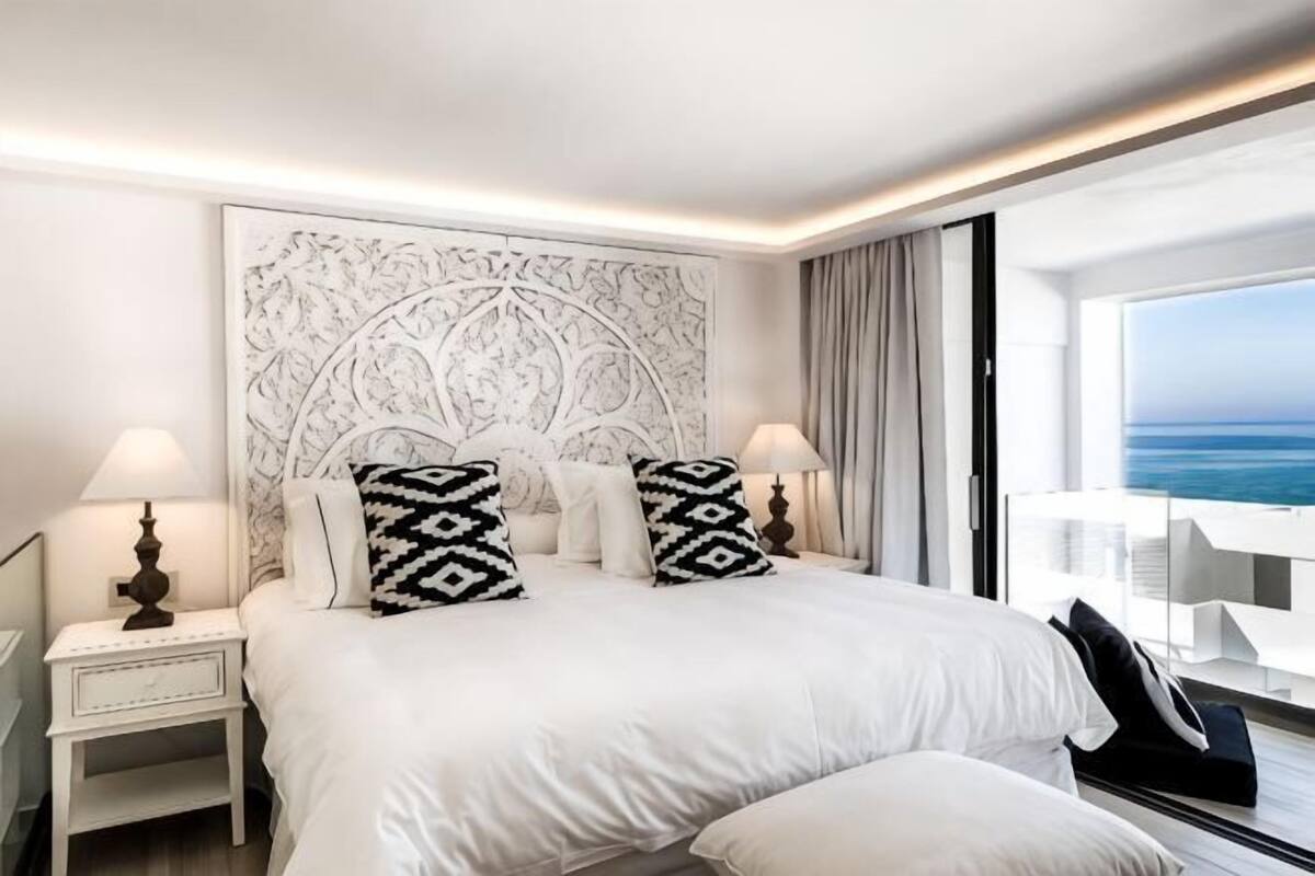 A hotel room with a large bed, two bedside tables with lamps, a decorative headboard, and a view of the ocean.