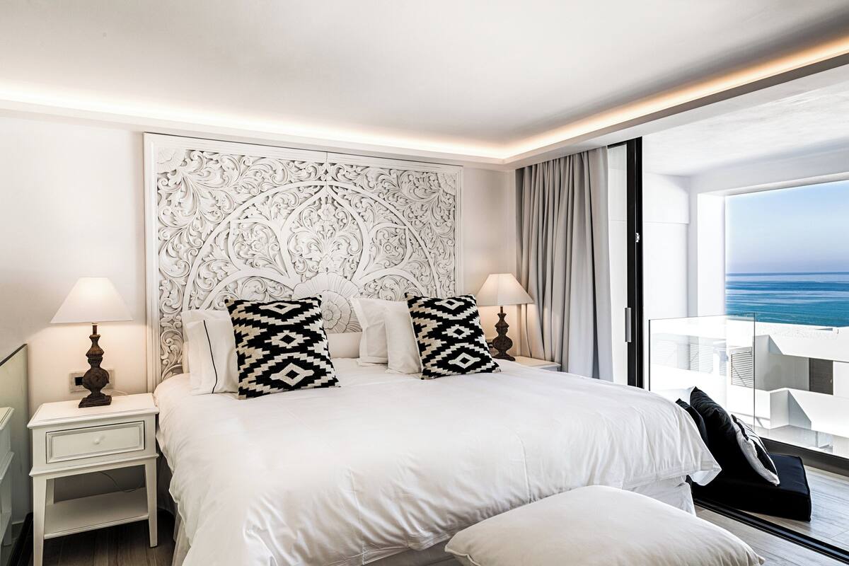 A modern hotel room with a large bed, two bedside lamps, a decorative wall panel, and a view of the ocean.