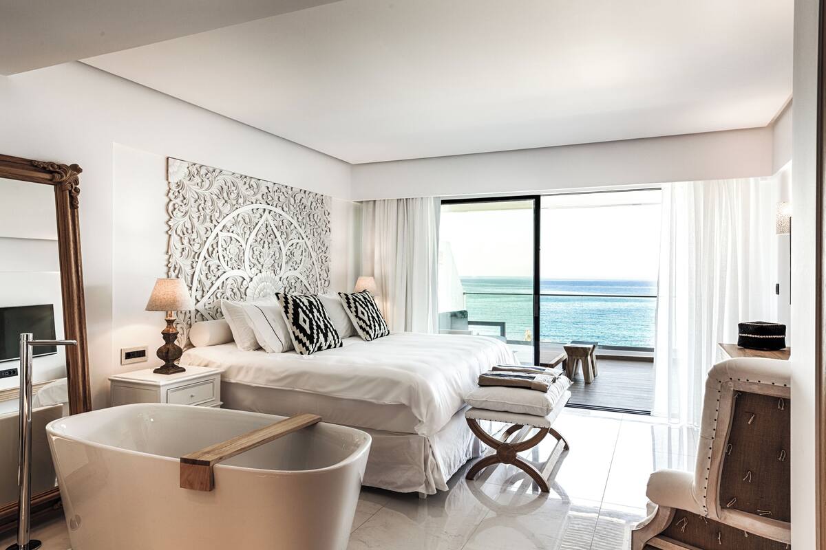 A modern hotel room with a large bed, a bathtub, a balcony with a view of the sea, and a large mirror.