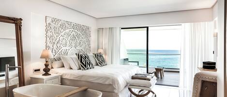A modern hotel room with a large bed, a bathtub, a balcony with a view of the sea, and a large mirror.