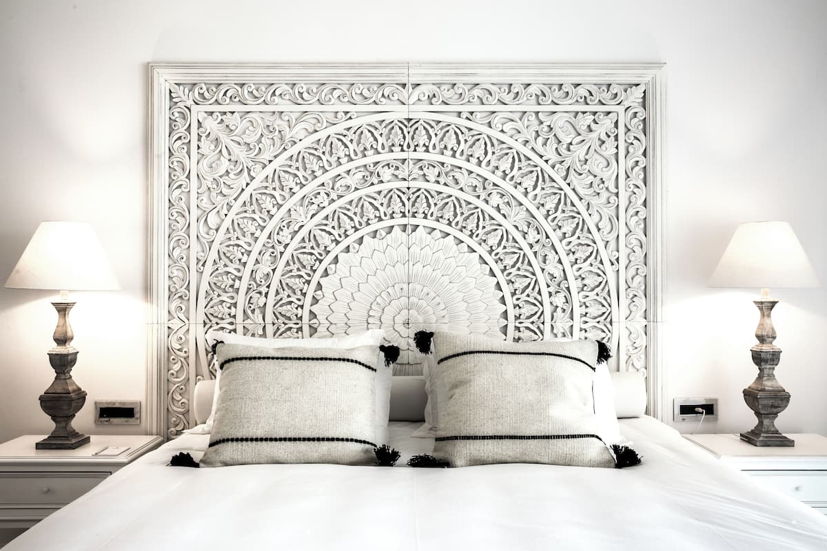 A neatly made bed with white bedding and two pillows, flanked by two bedside tables with lamps, against a wall with an ornate, symmetrical headboard.