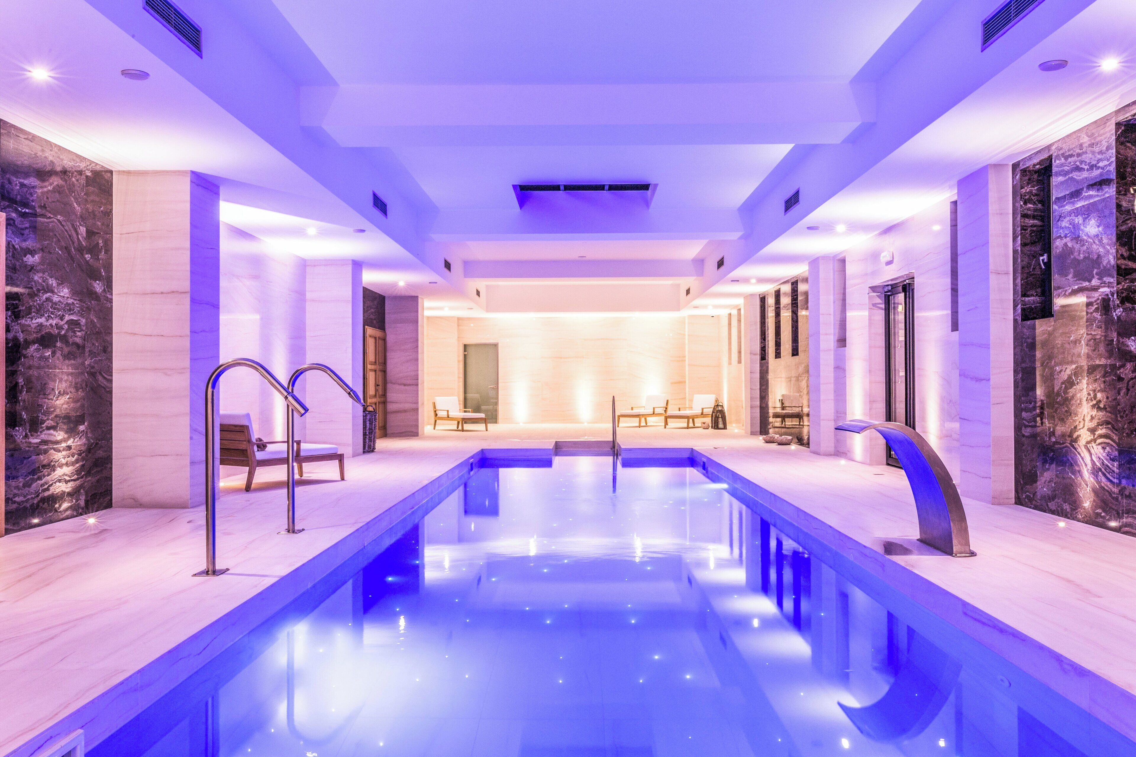 Piscina interior moderna com água azul cristalina, ladeada por paredes de mármore e equipada com vários pontos de acesso.