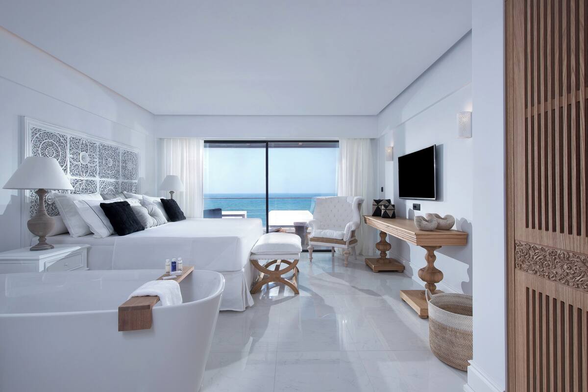 A modern hotel room with a large bed, a sitting area, a bathtub, and a view of the ocean.