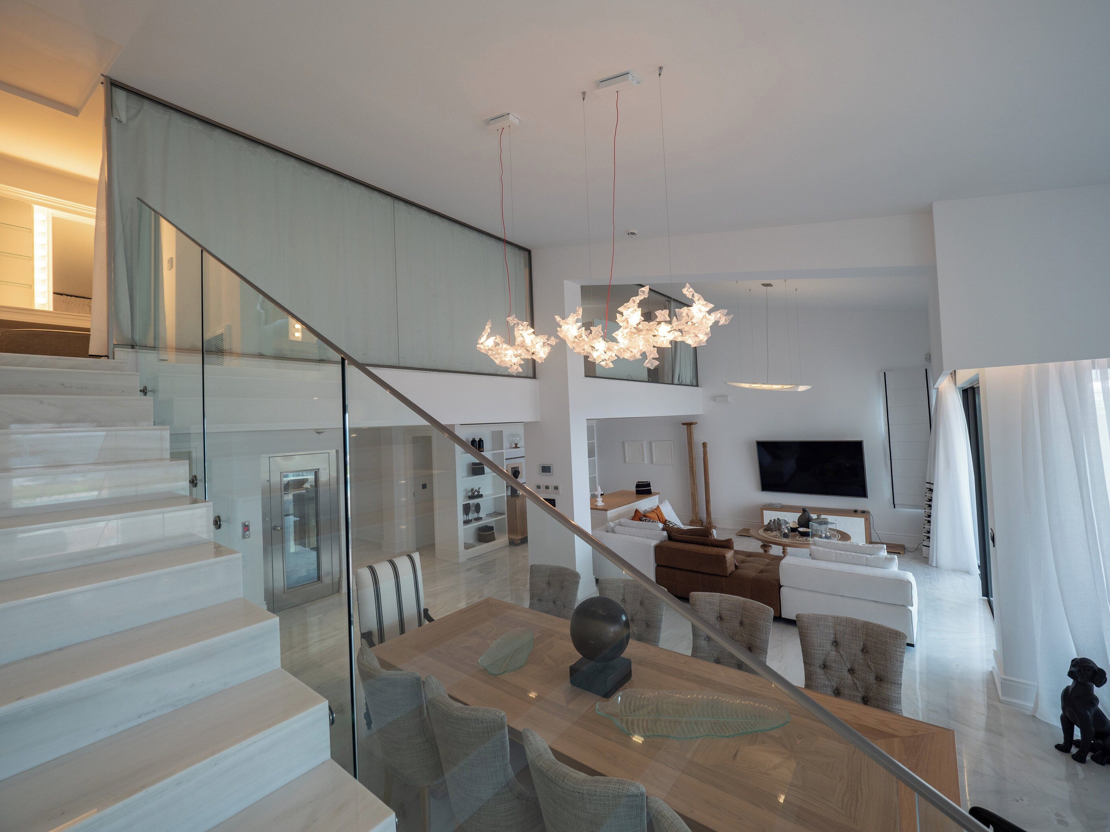 Una sala de estar moderna con una escalera de cristal, un comedor con mesa de cristal y un televisor al fondo.