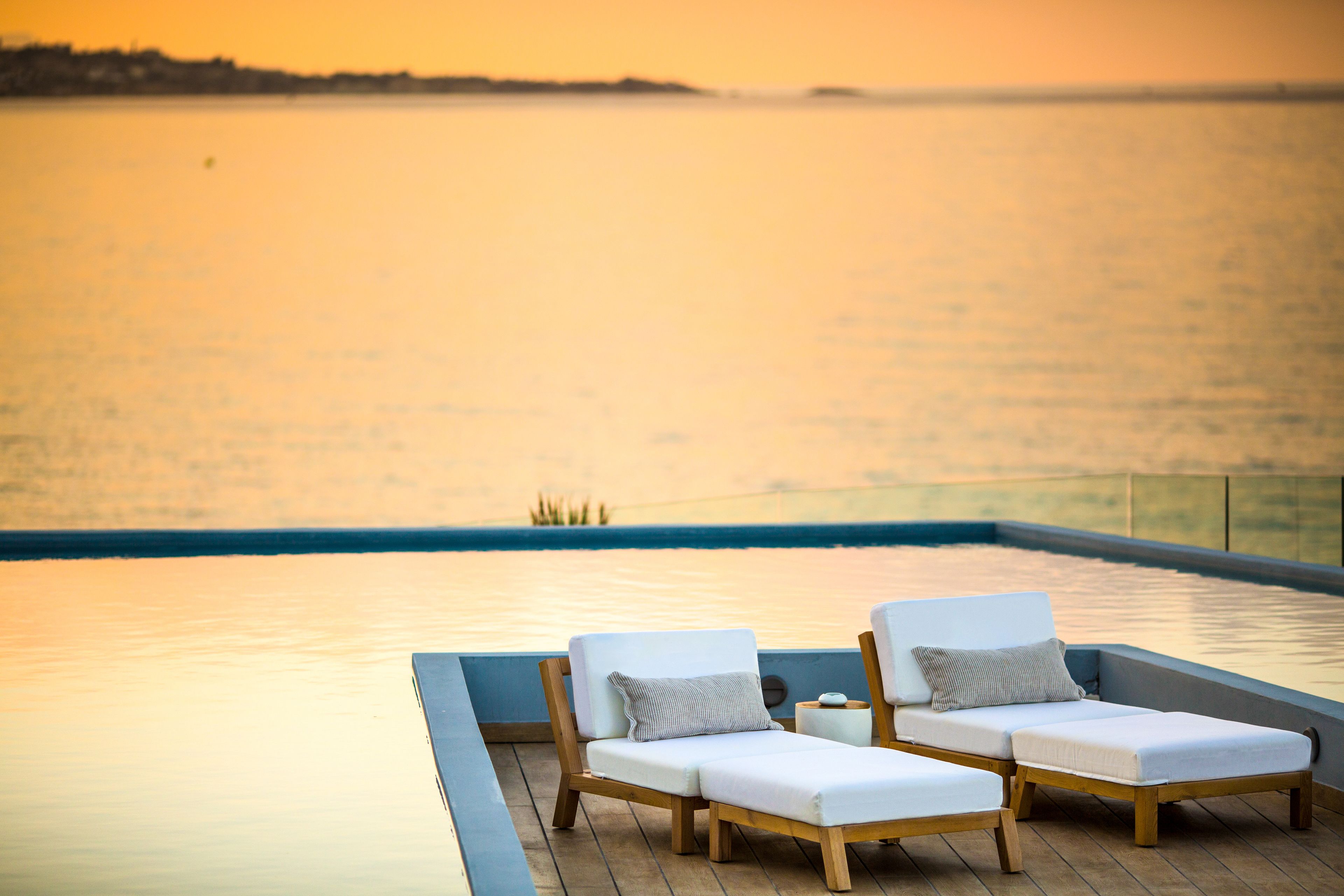 Un área junto a la piscina con dos sillones de descanso y una mesita, con vistas a una masa de agua tranquila.