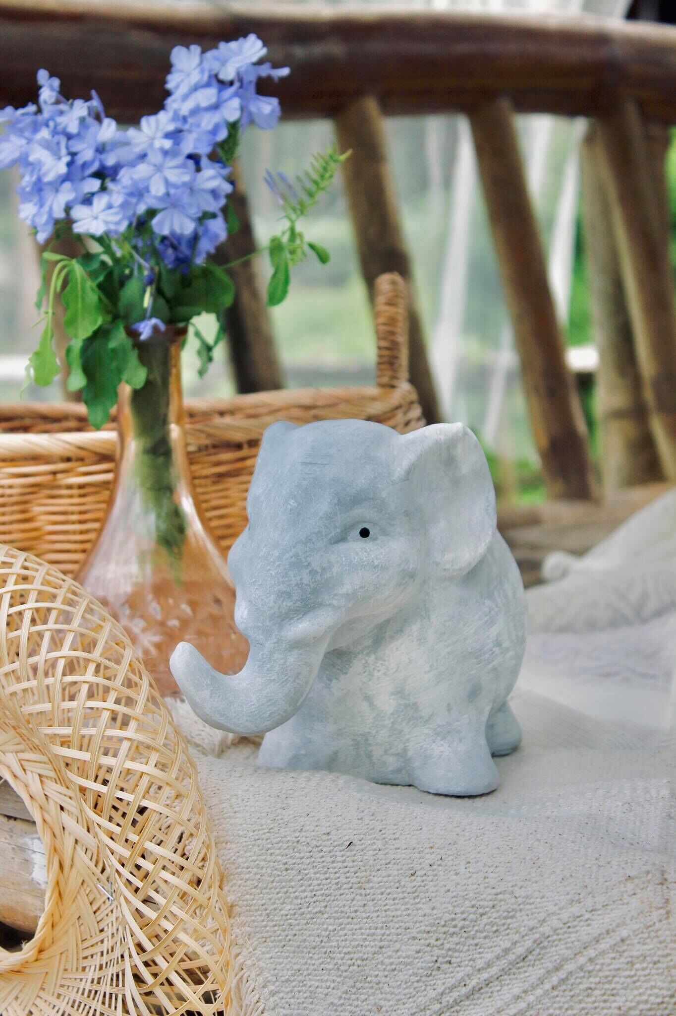A decorative elephant figurine on a textured surface, accompanied by a wicker basket and a vase with blue flowers.