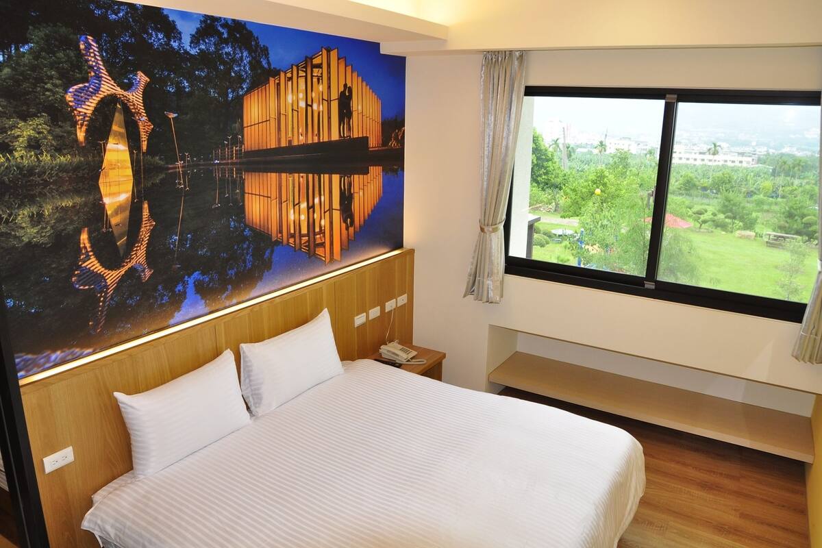 A hotel room with a large bed, a nightstand, a telephone, a window with a view of greenery and a building, and a mural on the wall.