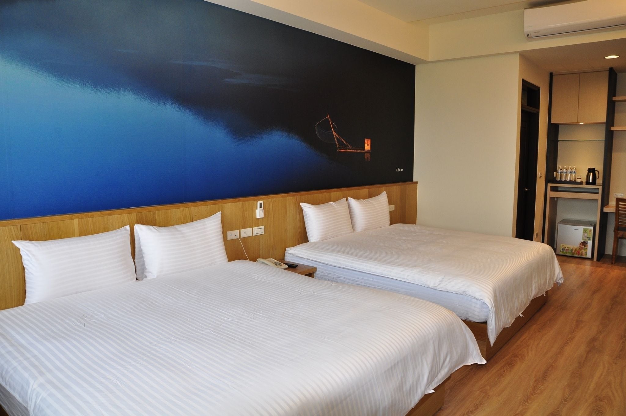 Hotellihuone, jossa on kaksi sänkyä, suuri seinämaalaus, puinen pääty ja pieni keittonurkkaus.