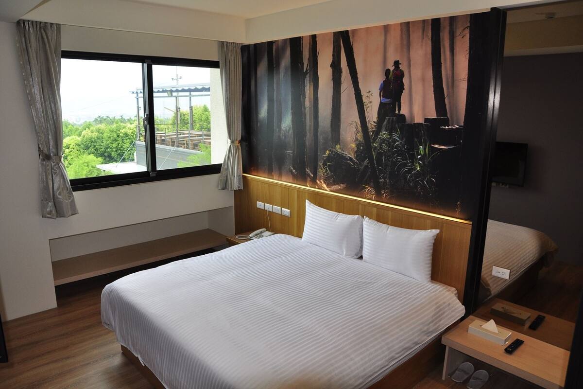 A hotel room with a large bed, a nightstand, a telephone, and a view of a forest through a window.