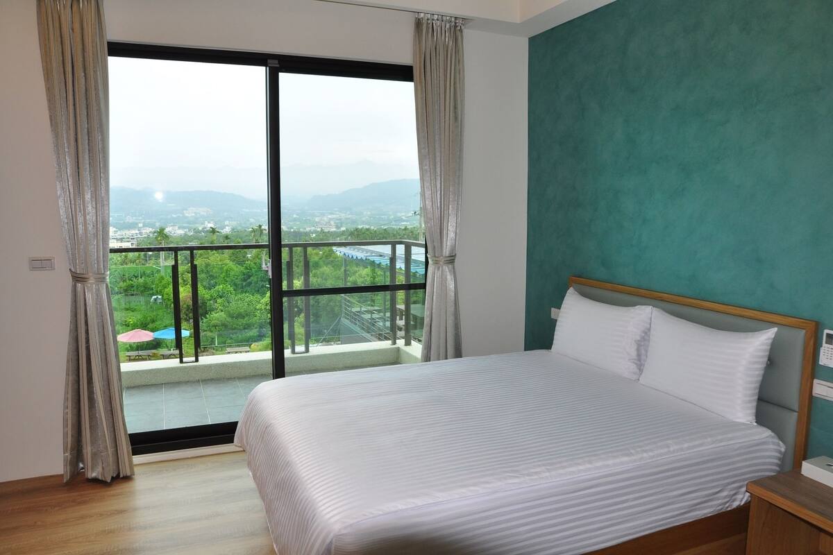 A bedroom with a large bed, a view of greenery and mountains, and a balcony with a railing.