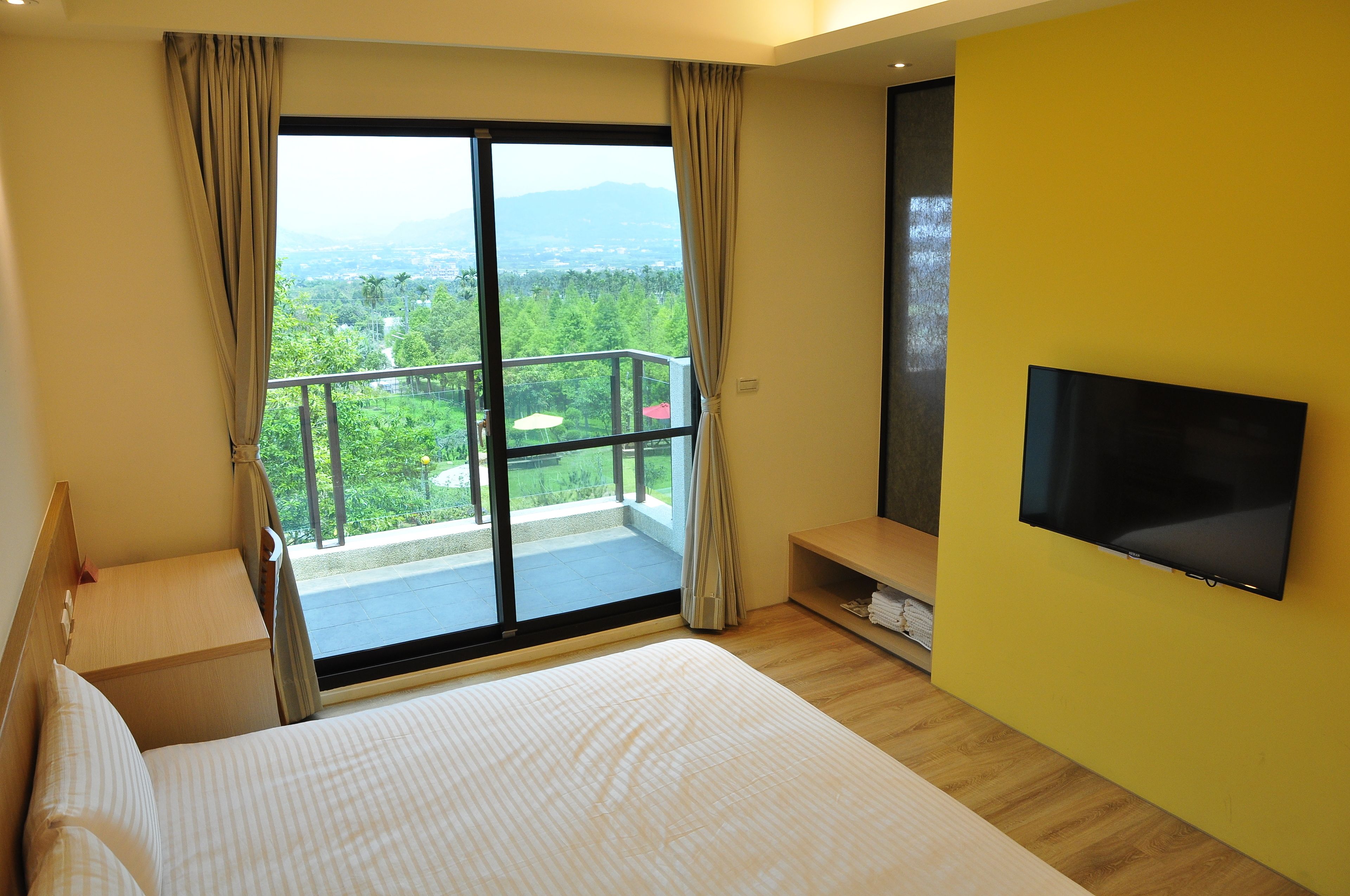 Een hotelkamer met een groot bed, een aan de muur bevestigde televisie, een balkon met uitzicht op groen en bergen, en een houten vloer.