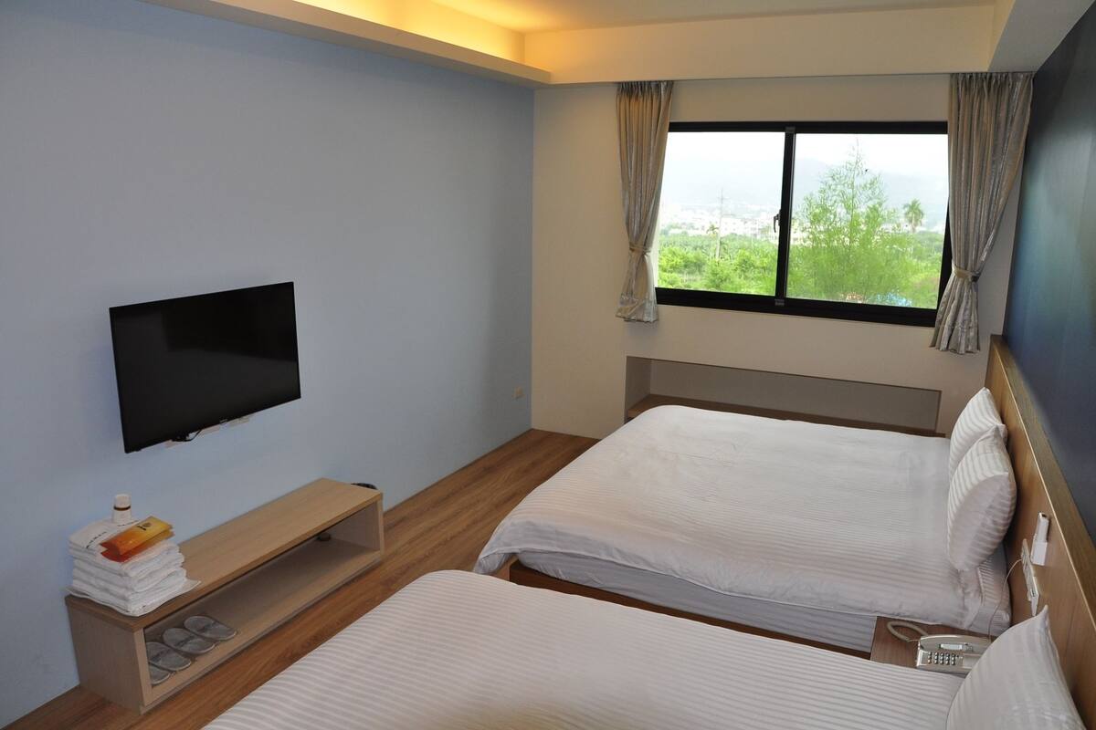 A hotel room with two beds, a TV mounted on the wall, a wooden bench with a small shelf, and a window with curtains.