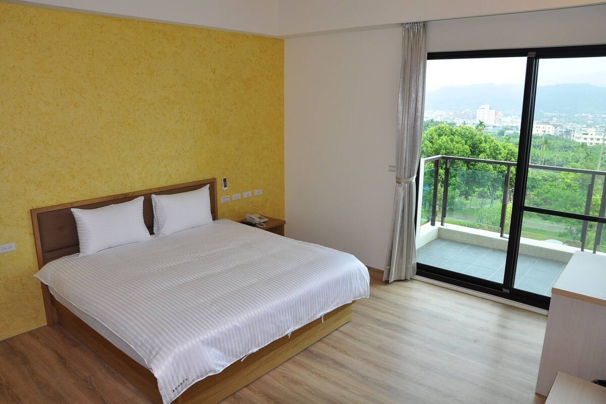 A hotel room with a bed, a nightstand, a phone, and a view of greenery and buildings through large windows.