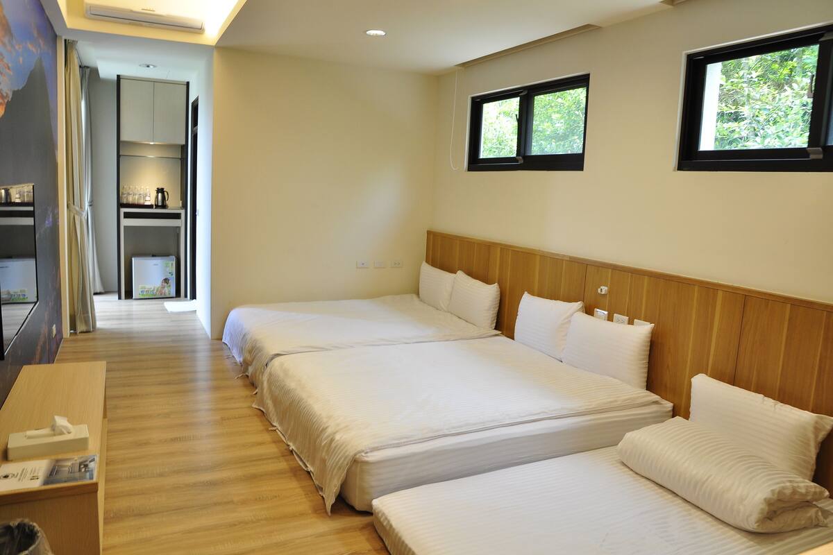 A hotel room with two beds, a wooden headboard, a small desk, and a view of greenery through the windows.