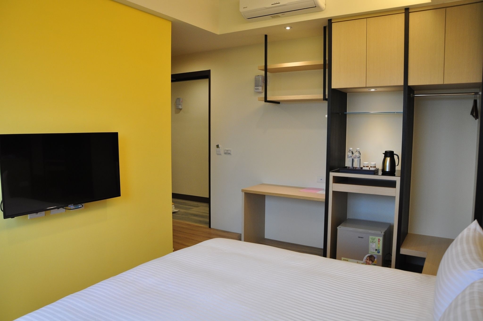 A hotel room with a bed, a flat-screen TV mounted on the wall, a desk with a coffee maker, and a small refrigerator.